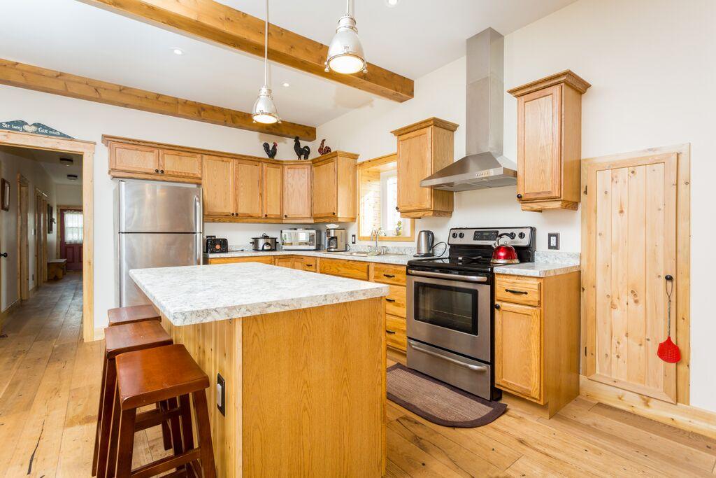 An open concept kitchen features wooden cabinets and granite countertops. Stainless steel appliances are visible, including a refrigerator and oven. An island with seating for three is located in the center, while wooden flooring and exposed ceiling beams add character to the space.