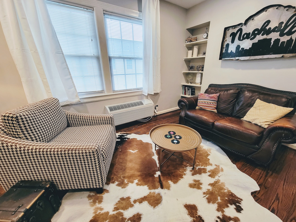 A cozy living area features a checker-patterned armchair and a leather sofa, complementing a round coffee table atop a patterned area rug. Natural light filters through large windows adorned with light curtains, creating a relaxed ambiance.