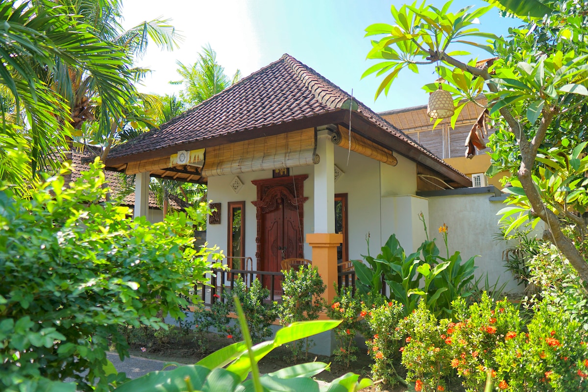 A charming bungalow is surrounded by lush tropical greenery and vibrant flowers. The thatched roof complements the wooden door, and large windows allow natural light to brighten the interior. A pathway leads through a well-maintained garden, inviting relaxation in a serene environment.