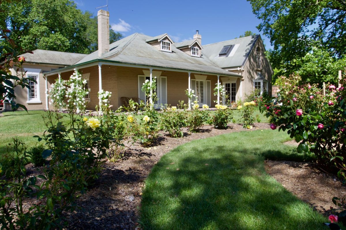 A charming homestead is surrounded by lush gardens filled with blooming roses and greenery. The exterior features a sloped roof and multiple windows, allowing natural light to illuminate the space. A winding path leads through the manicured lawn, enhancing the tranquil country atmosphere.