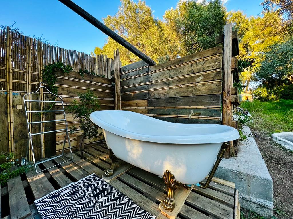 An outdoor bathing area features a vintage freestanding tub set on wooden pallets. Surrounding the tub is a rustic wooden fence adorned with greenery, enhancing the natural ambiance. A patterned rug lies beneath, adding comfort to the space.