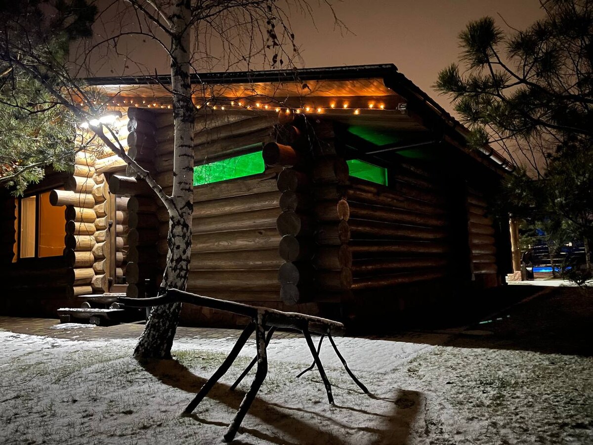 The exterior of a wooden cabin, designed in a Swiss style, is highlighted by soft green light emanating from the windows. The cabin is surrounded by snow and trees, creating a serene winter atmosphere. Decorative lighting adds a warm touch to the rustic structure.