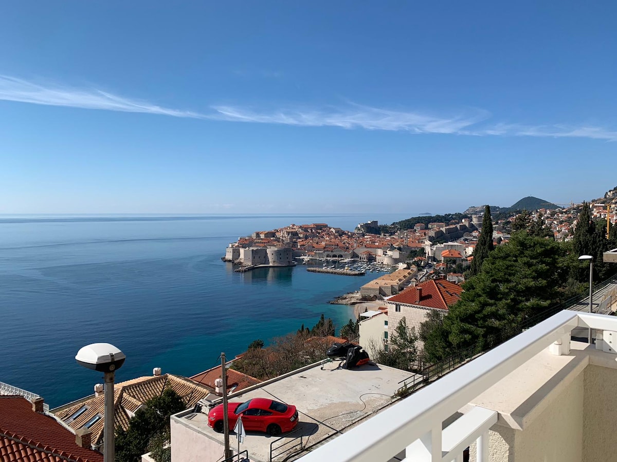 A panoramic view of Dubrovnik's old town and the island of Lokrum is displayed, featuring vibrant rooftops and serene turquoise waters. The clear sky complements the scenic landscape, while a modern railing frames the foreground, offering an inviting perspective of the captivating surroundings.