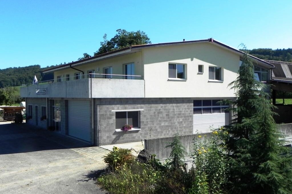 A two-story building is displayed with a light-colored facade and large windows. A balcony extends from the upper level, presenting an outdoor space. Surrounding greenery is visible, enhancing the natural setting of the location.
