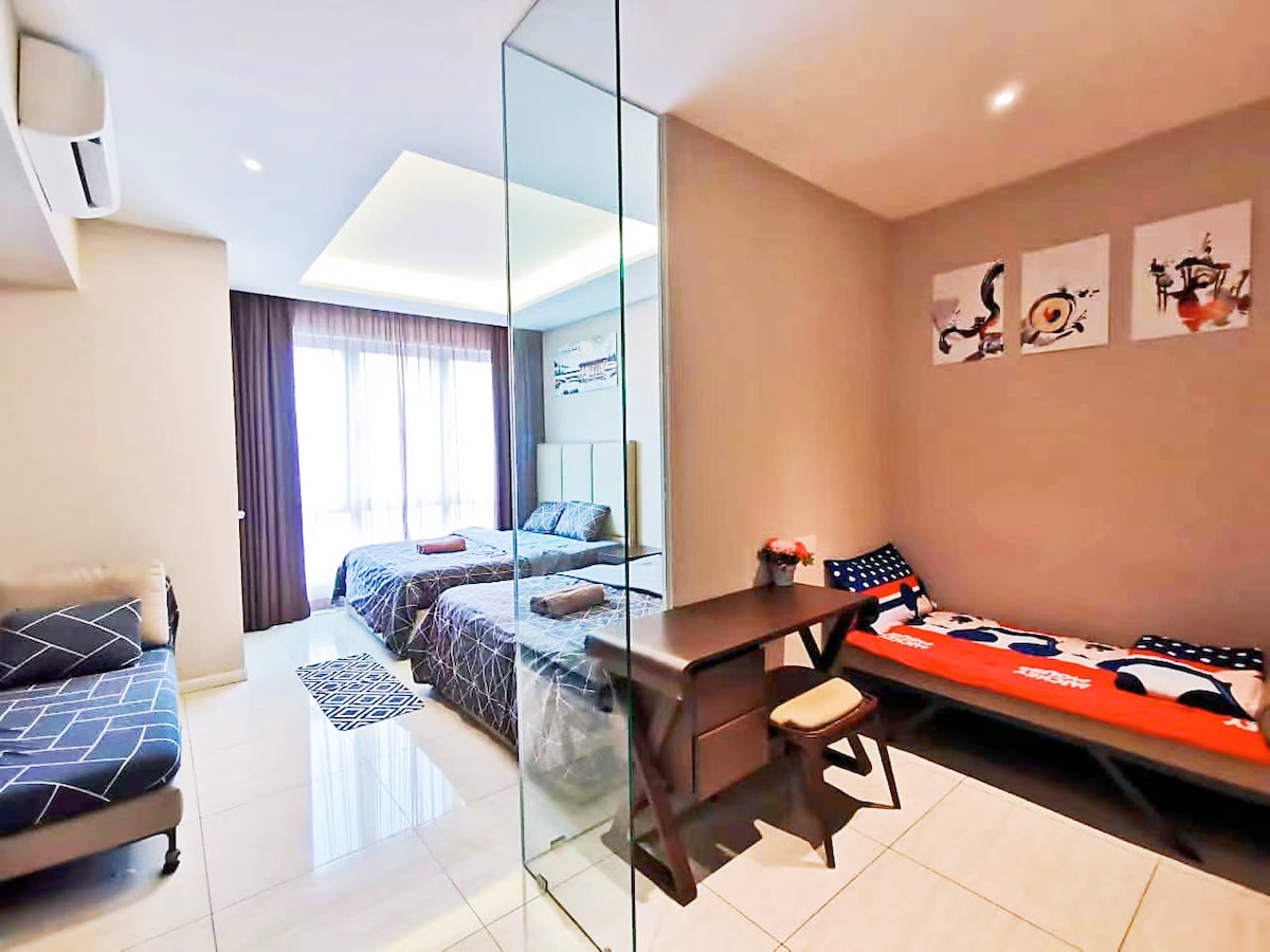 A spacious room features two queen-size beds covered with comforters, alongside a three-seater sofa. A working desk is present next to a single bed with a bolster. Large windows provide natural light, complemented by modern decor and a neutral color palette.