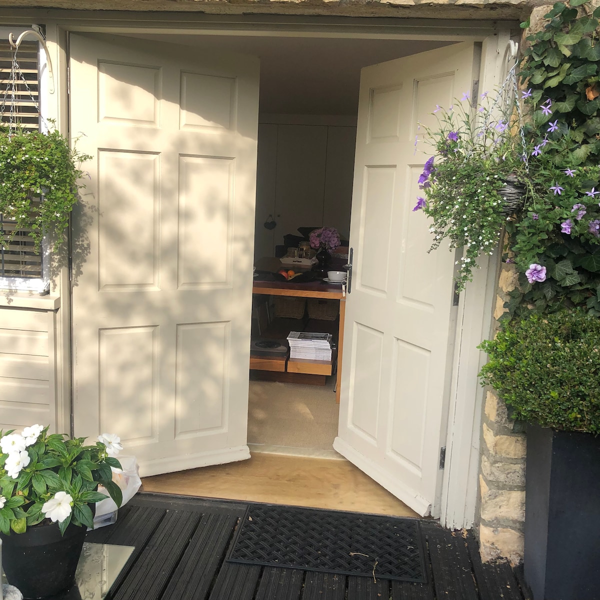 The image shows a set of double doors that are open, leading into a bright studio space. Flowering plants hang from the doors, and a potted plant is visible on the floor, adding a touch of greenery. The interior appears inviting and well-lit.