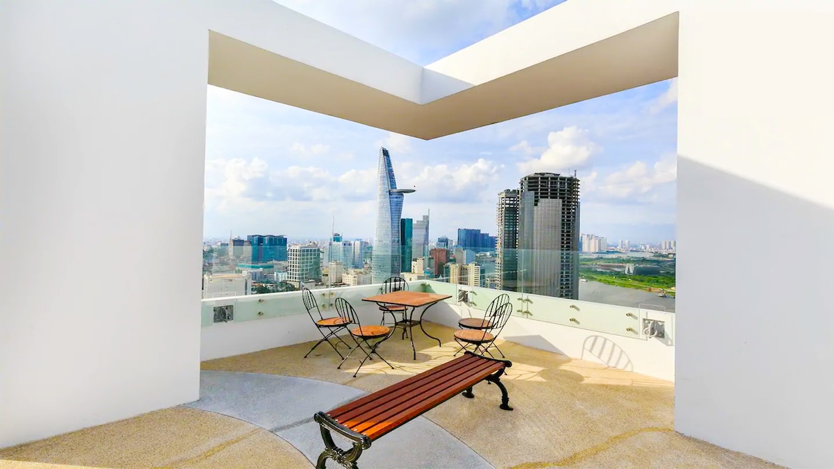 An outdoor seating area features a round table with four accompanying chairs, positioned against a backdrop of city skyscrapers. A bench complements the setting, offering a view of the skyline and the river, framed by sleek white architectural details.