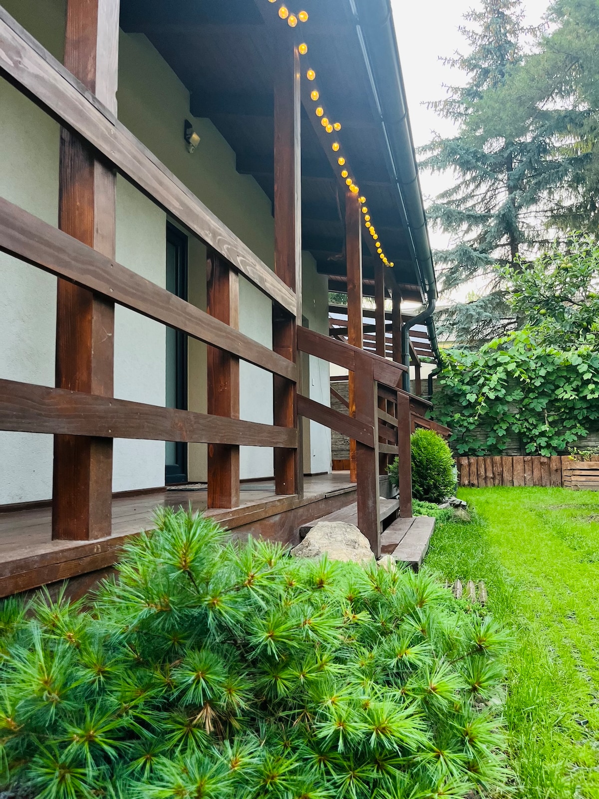 A wooden deck is framed by a sturdy railing, leading to a peaceful garden area adorned with neatly arranged greenery. Soft lighting is strung above, enhancing the outdoor space, while a natural backdrop of trees and shrubs is visible in the background.