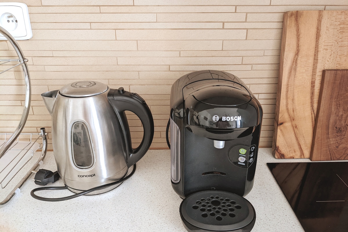 A sleek stainless steel kettle is positioned next to a modern black coffee machine, both set against a light, textured backsplash. The clean countertop features a minimalistic design, enhancing the functional kitchen space.
