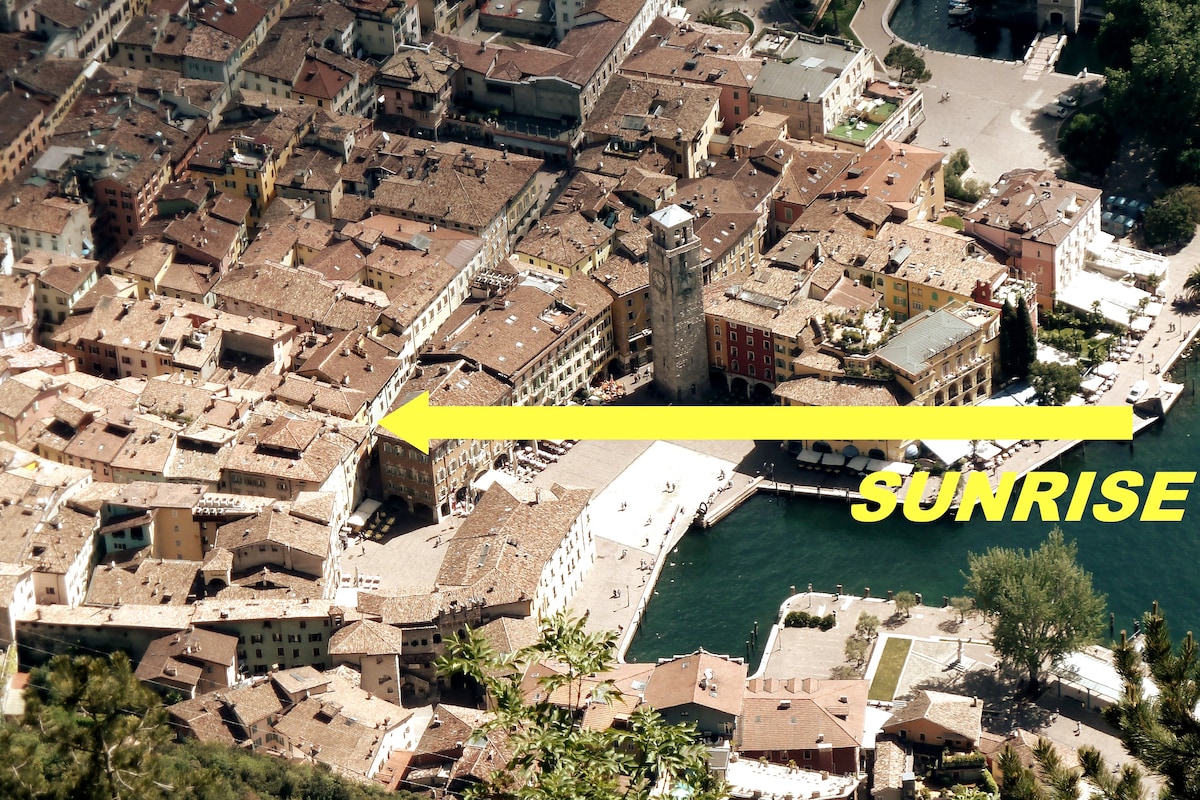 An aerial view captures the historic center of Riva del Garda, showcasing terracotta rooftops and narrow alleyways. The edge of the lake is visible, along with the Apponale Tower. A bright yellow arrow emphasizes the direction of sunrise over the horizon.