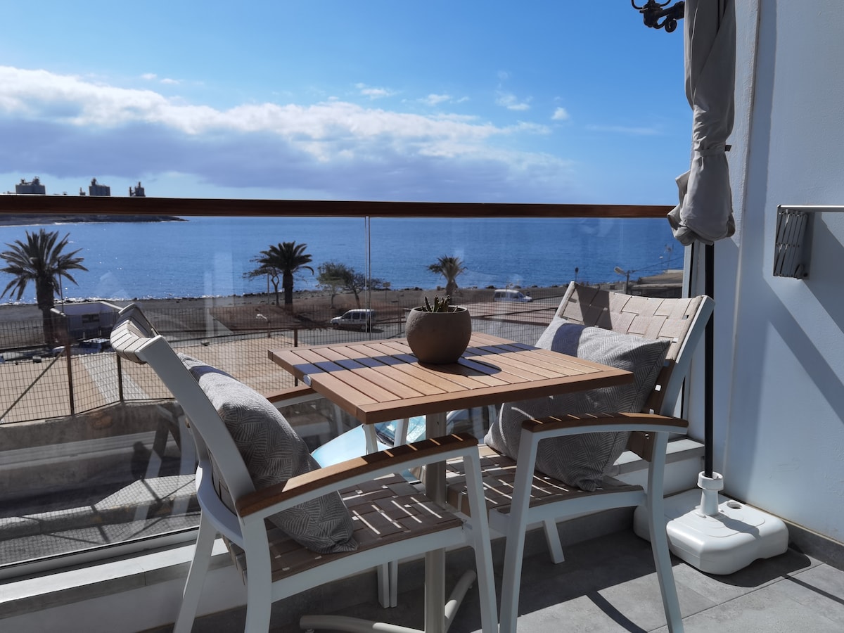 A private balcony features a small wooden table and two white chairs, complemented by soft cushions. A potted plant sits at the center of the table. The view includes the Atlantic Ocean and palm trees, with clear blue skies enhancing the coastal atmosphere.
