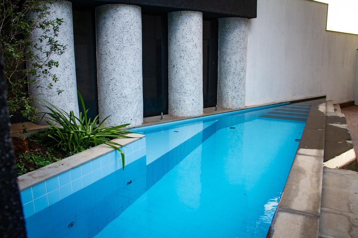 A serene swimming pool is framed by textured columns and complemented by greenery. The water reflects a clear blue, while the pool's smooth tiles enhance the clean lines of the setting. Natural light illuminates the area, creating a calm and refreshing atmosphere.
