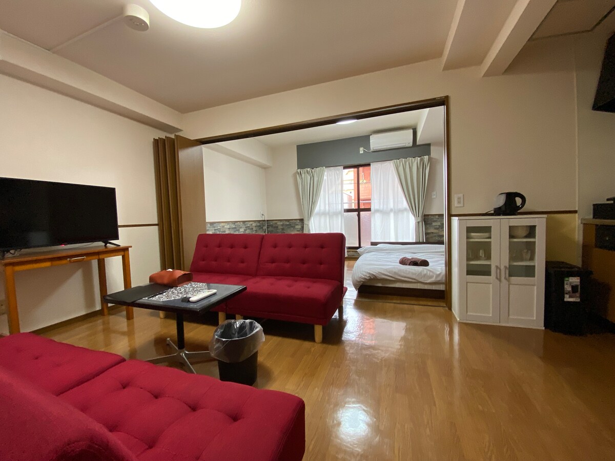The spacious living area features a red seating arrangement with two sofas, a black coffee table, and a television. Natural light enters through the curtains covering the window, illuminating the adjoining bedroom with a white bed and soft linens.