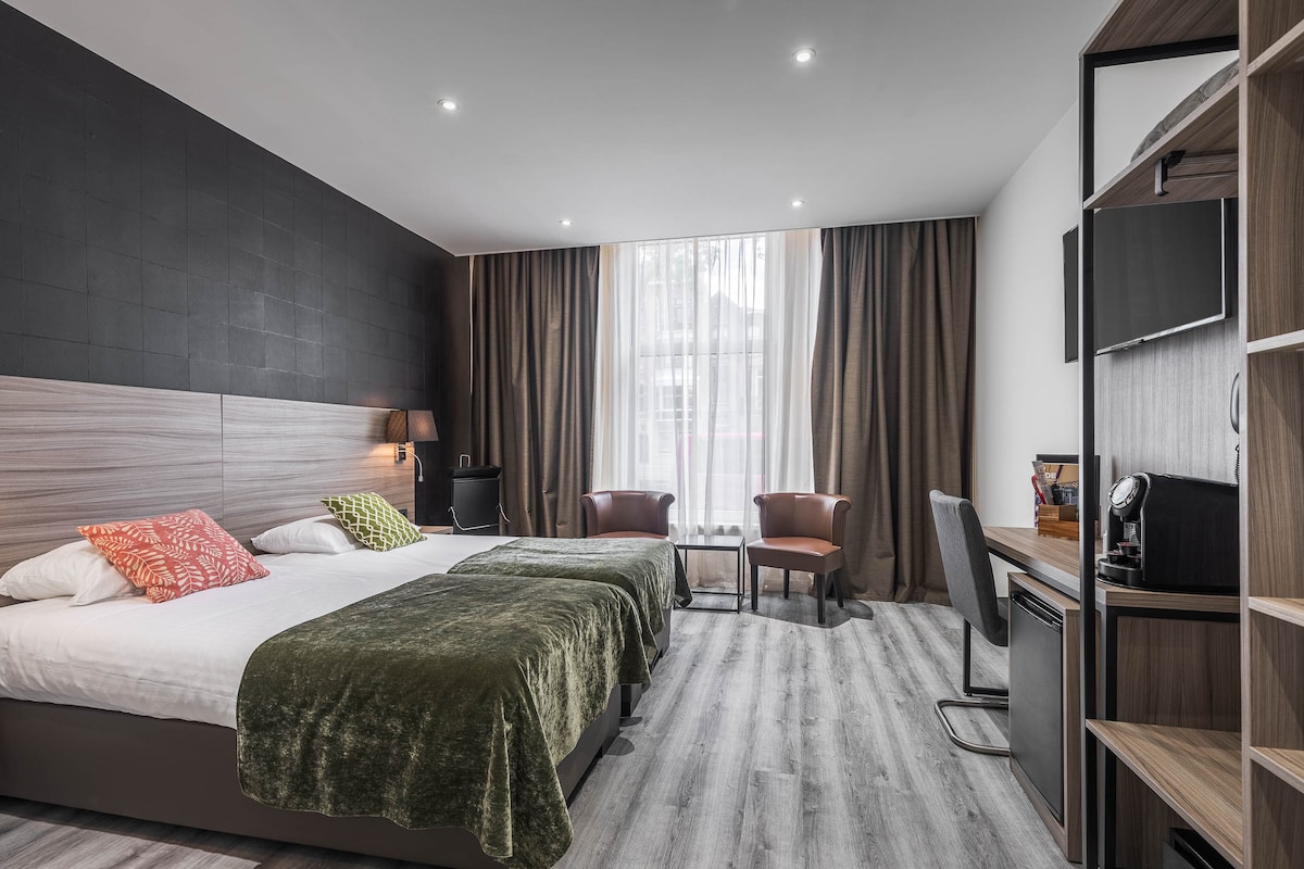 The room features two spacious twin beds topped with olive green throws. Natural light filters through large curtains, illuminating the space. A desk with a chair and a small seating area with two armchairs are arranged across from the bed, enhancing convenience.