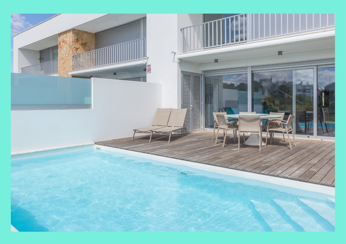 The outdoor area features a refreshing private pool surrounded by a wooden deck. The space includes sun loungers and a dining table with chairs, inviting relaxation and outdoor dining experiences. Large glass doors offer views of the interior, blending the spaces.