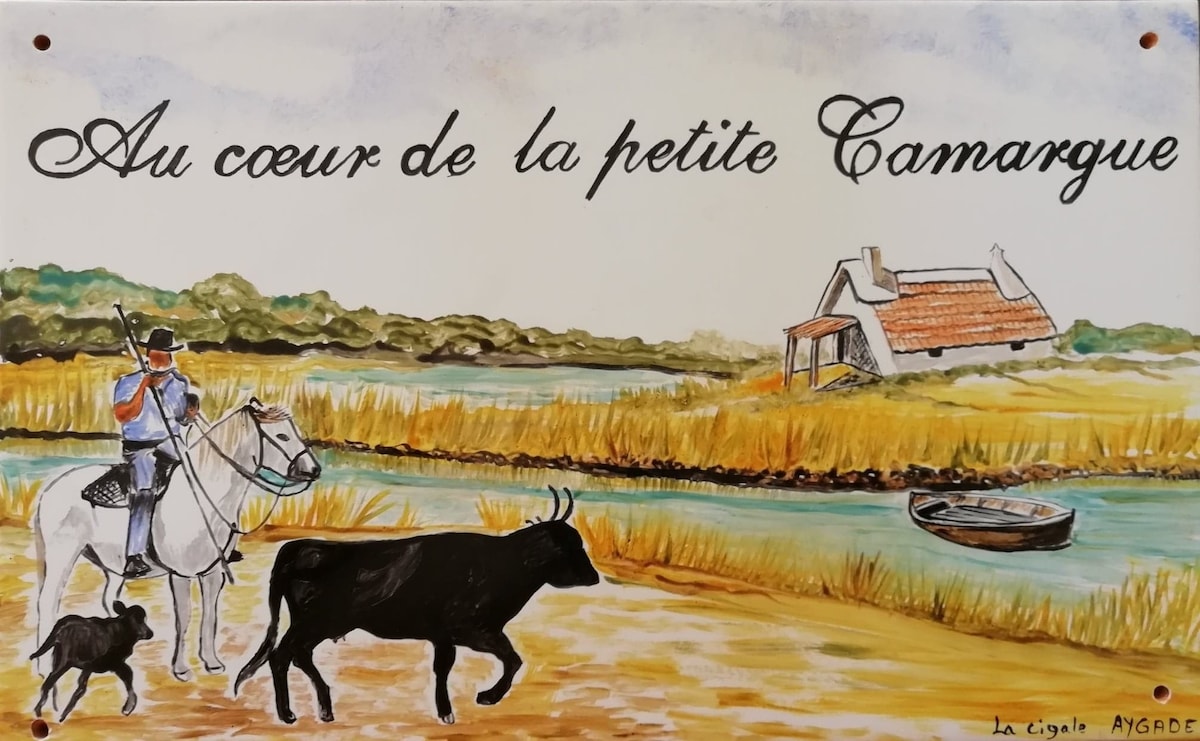 A decorative sign features the phrase 'Au coeur de la petite Camargue,' accompanied by an illustration of a mounted rider on a white horse. In the foreground, a black bull stands near a river, while a small boat is visible on the water, complemented by a rustic home in the background.