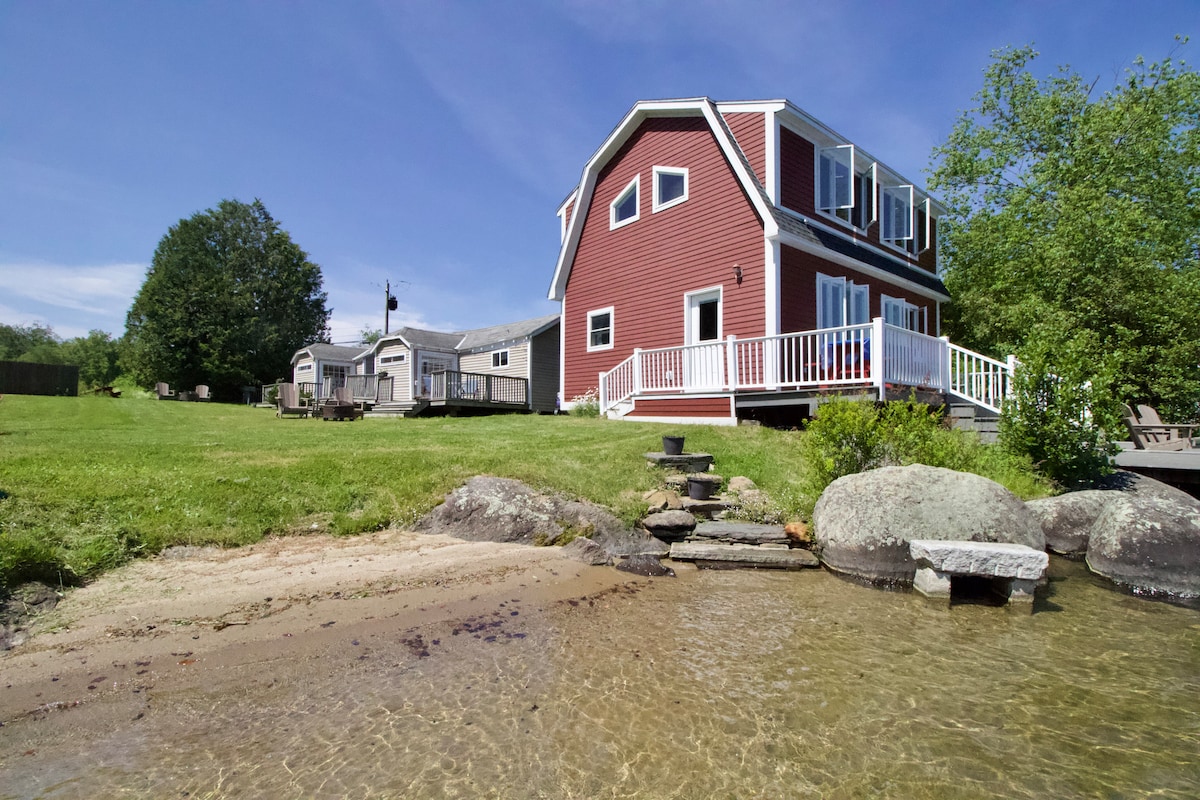A red cottage is set against a clear blue sky, featuring a white railing on the deck. Steps lead down to the sandy beach and crystal clear water, with grassy areas and trees enhancing the natural setting.
