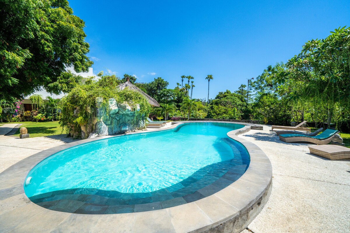 A large, curved swimming pool reflects the clear blue sky, surrounded by a spacious deck and tropical foliage. Comfortable lounge chairs are positioned nearby, and a thatched-roof gazebo can be glimpsed among the greenery, providing shade and a serene retreat.