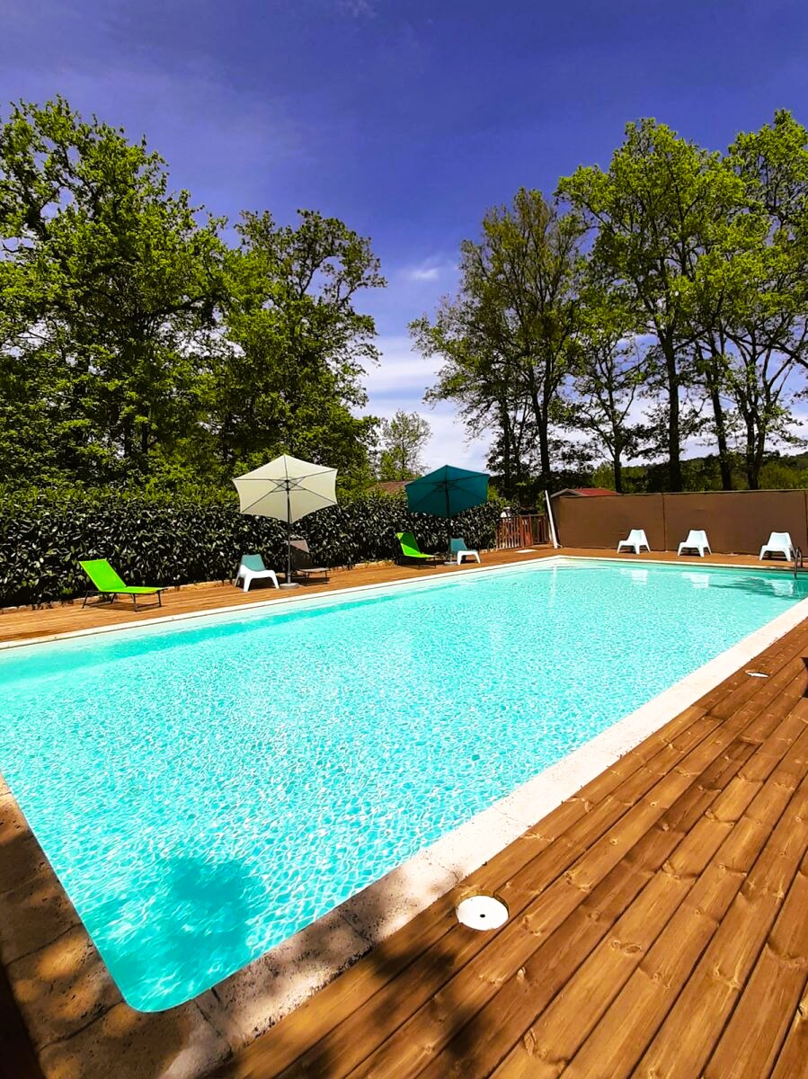 The outdoor heated pool is framed by a wooden deck, featuring several lounge chairs and umbrellas for shade. Surrounding greenery enhances the tranquil setting, providing a peaceful retreat in a sunny environment.