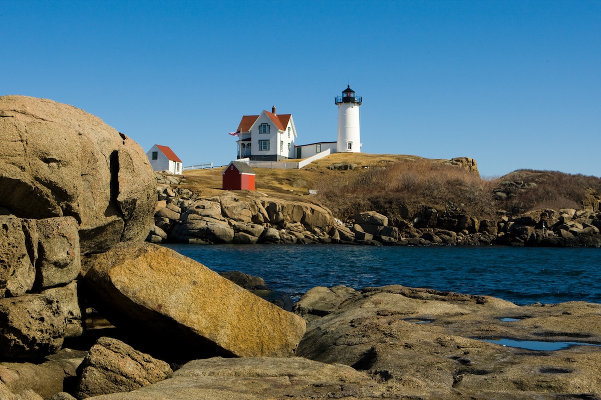 A historic lighthouse sits atop a rocky outcrop, framed by a clear blue sky. The white structure features a dark lantern room and a red-roofed keeper's house, while surrounding boulders and gentle waves add to the coastal scenery.