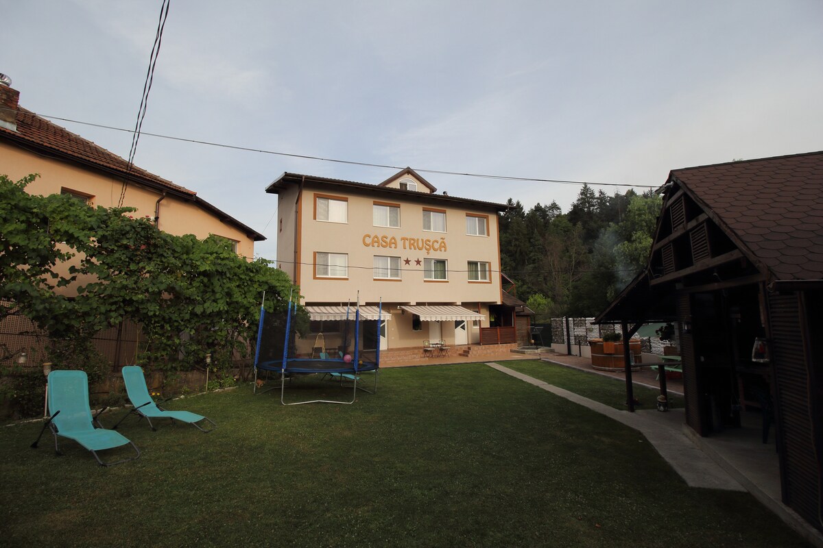 A two-story building named 'Casa Trusca' is set in a grassy yard. Several lounge chairs are positioned on the lawn. A trampoline can be seen on one side, while an outdoor grill and shaded gazebo are visible on the other side.