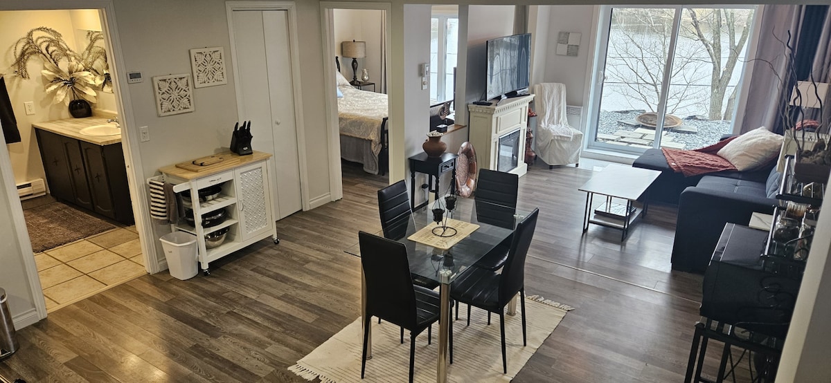 An open-concept living area is displayed, featuring a dining table with chairs and a view of large windows overlooking the water. A cozy sofa is positioned near a television, and a bedroom area can be seen through a doorway.