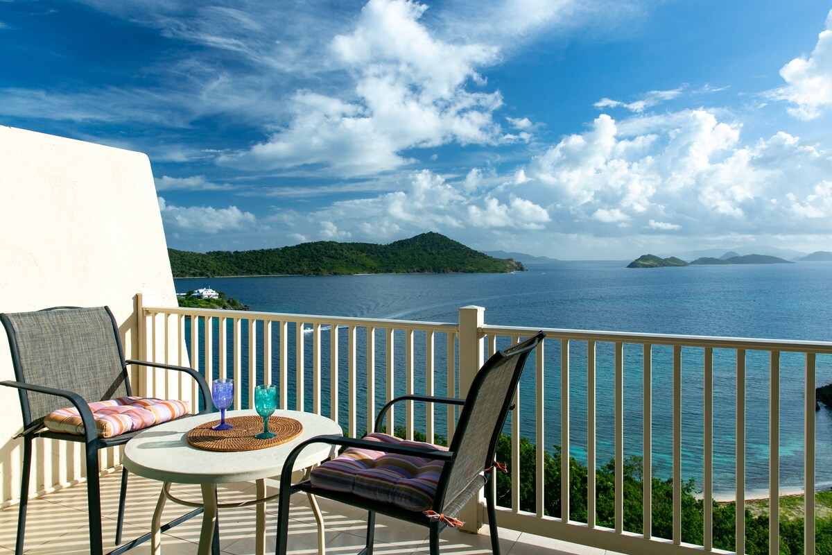 A private balcony features two chairs and a circular table, offering a serene spot to enjoy the stunning views of the Caribbean Sea. The horizon showcases distant islands under a clear blue sky, accentuated by scattered clouds.