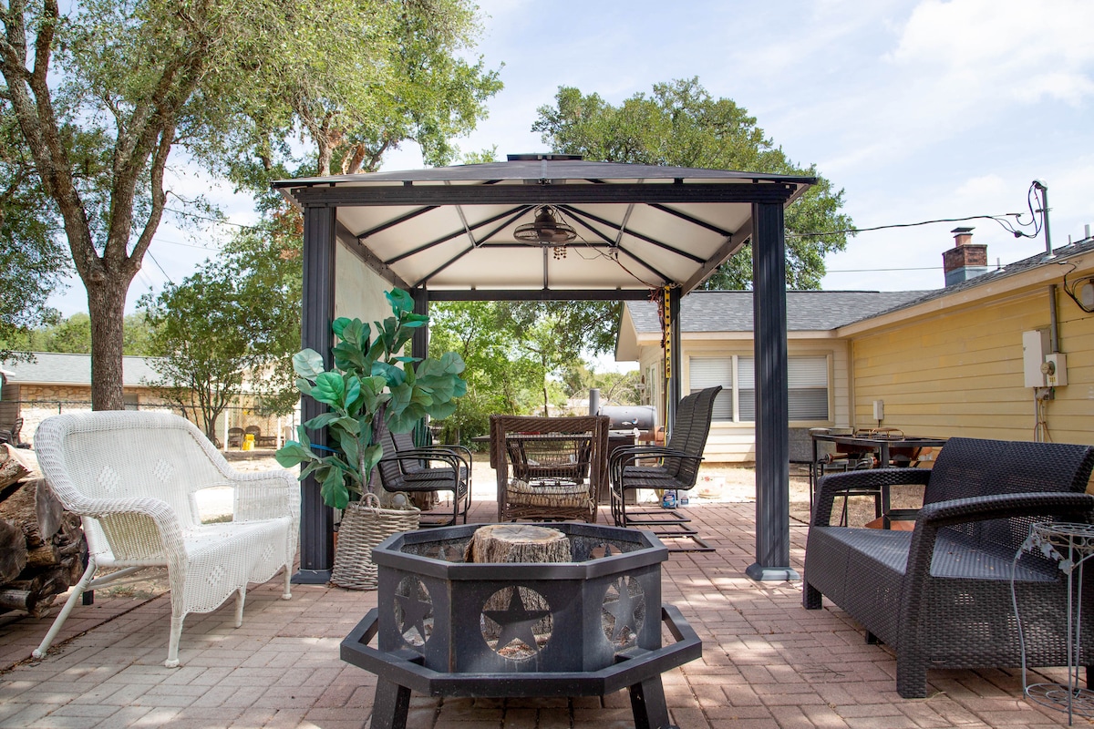 An outdoor seating area is provided under a covered gazebo, featuring a mix of wicker and metal chairs arranged around a circular fire pit. Lush green plants add a touch of nature, while the surrounding trees offer shade and a relaxed atmosphere.