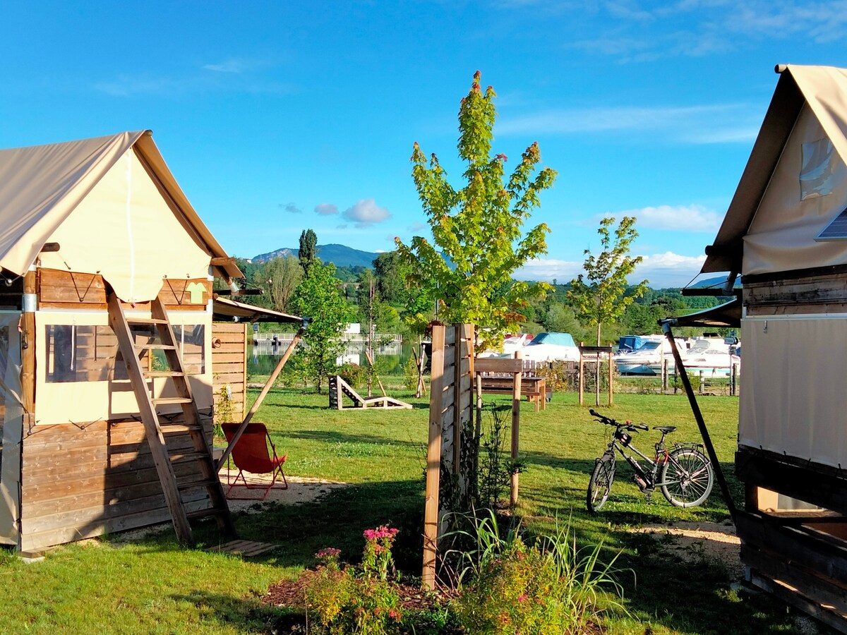 Two tents-lodges are situated amidst a green landscape, with a tranquil view of the water and distant mountains. A small seating area is visible beside one tent, while a bicycle is parked nearby, suggesting a convenient access to cycling paths.