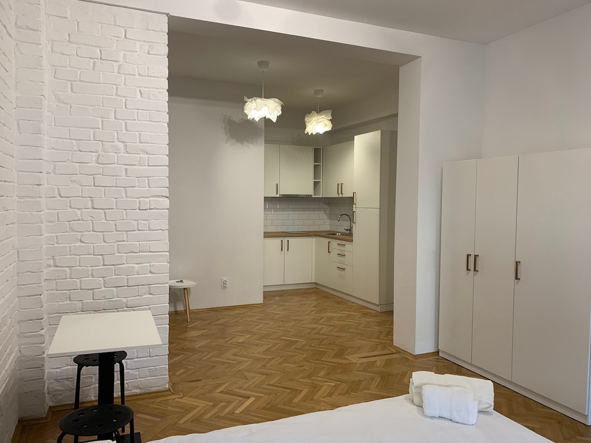 A spacious studio features light wood flooring and white brick walls. The kitchenette includes modern cabinetry, with light fixtures complementing the bright, airy atmosphere. A small dining table and minimalist chair are positioned nearby, while storage cabinets offer practical space for belongings.