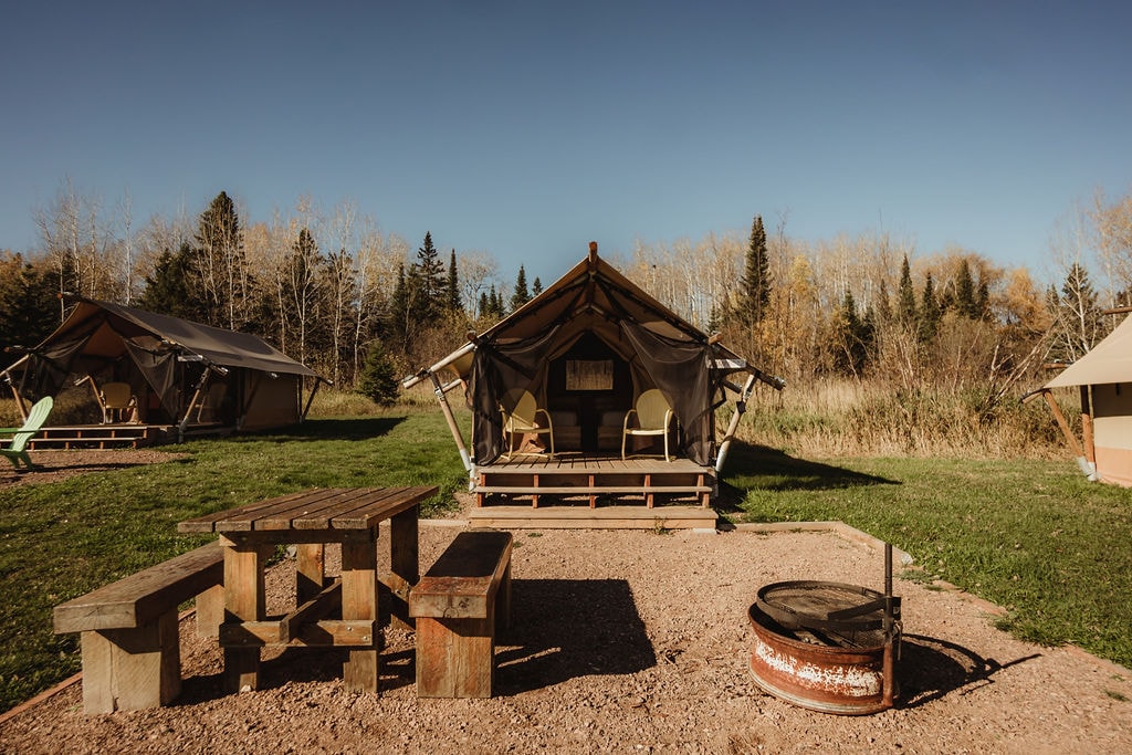 Canvas wall tents are set on a natural landscape, surrounded by tall grasses and trees. A picnic table made of wood and a fire ring are positioned in front of the tents, offering a space for outdoor gatherings. Two armchairs are placed on the deck of one tent.