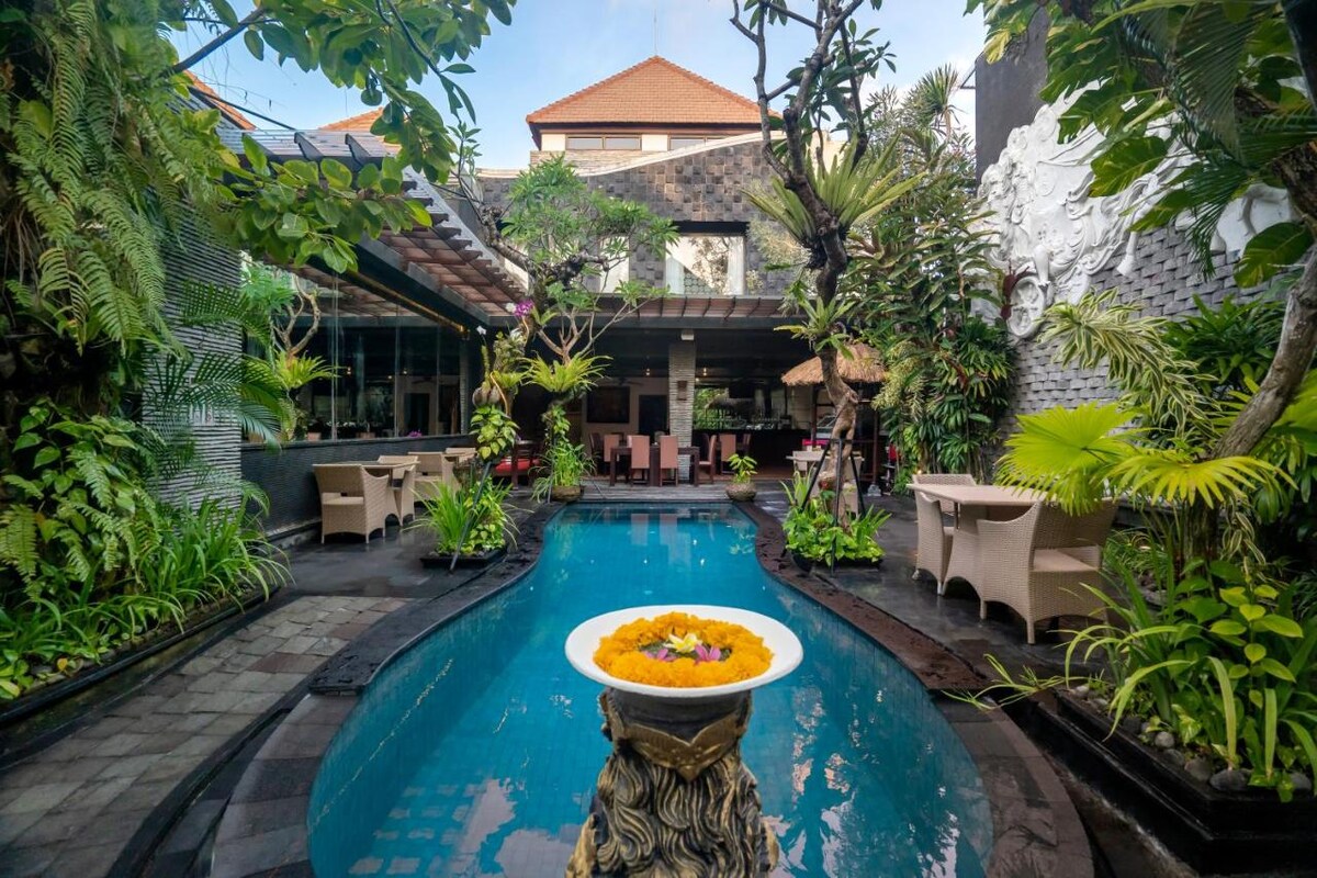 A serene private pool, surrounded by lush tropical greenery, features a decorative stone fountain at its center. Comfortable seating areas with light-colored chairs are positioned alongside the water, enhancing the tranquil setting. The backdrop showcases modern architecture amid vibrant plants.