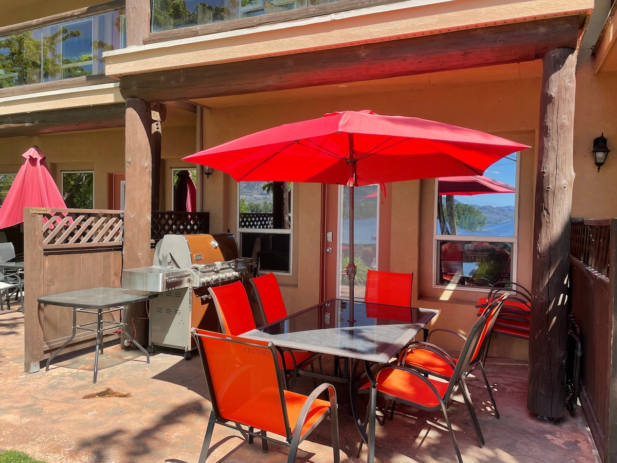An inviting outdoor patio area is furnished with a table and chairs under a vibrant red umbrella. A natural gas BBQ is positioned nearby, with a view of the lake visible through the adjacent windows.