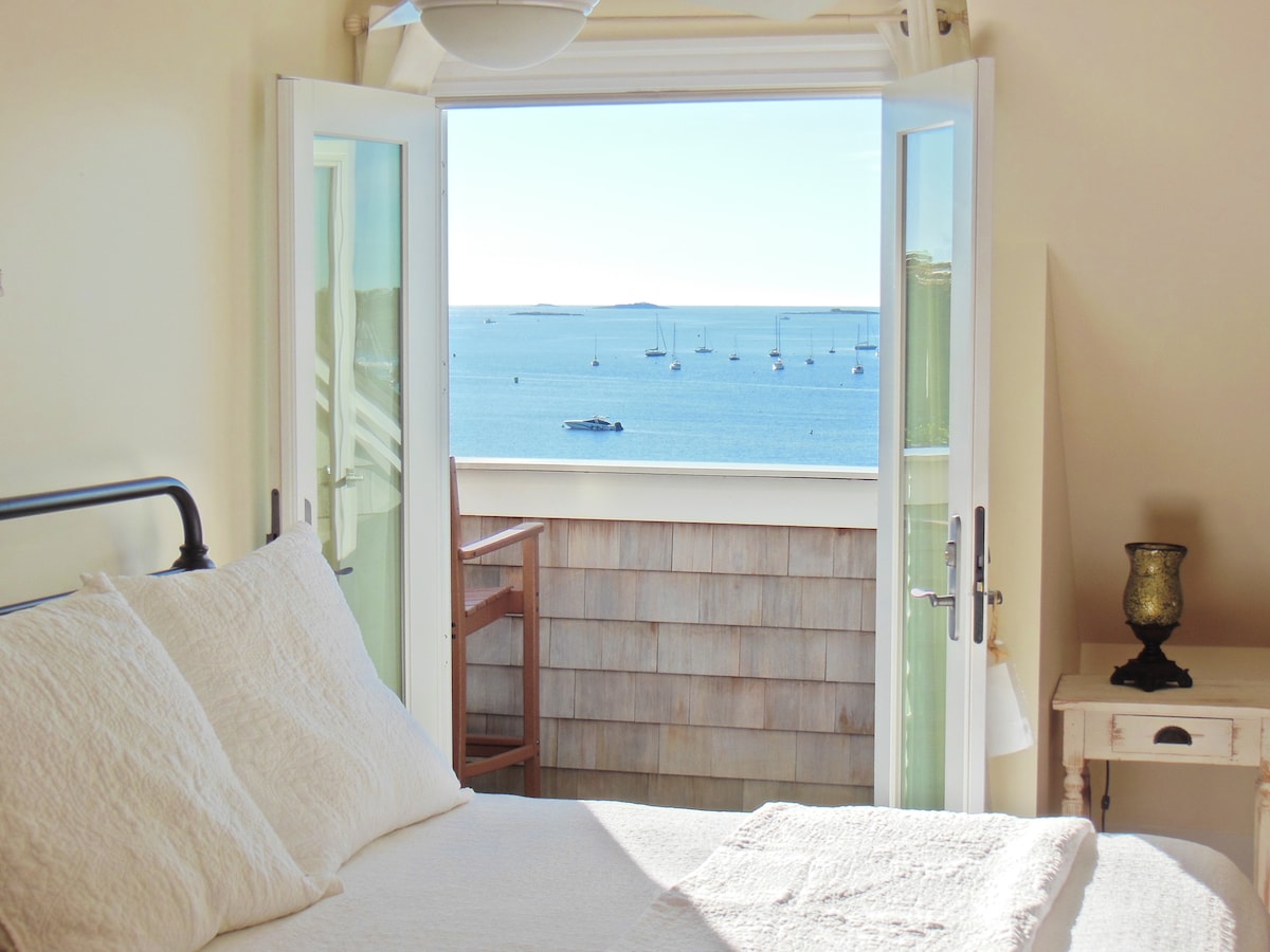 A bright bedroom features large open doors leading to an oceanfront balcony. The spacious bed is adorned with light bedding. Views of the water display boats gently bobbing, complemented by the soft hues of the open sky.