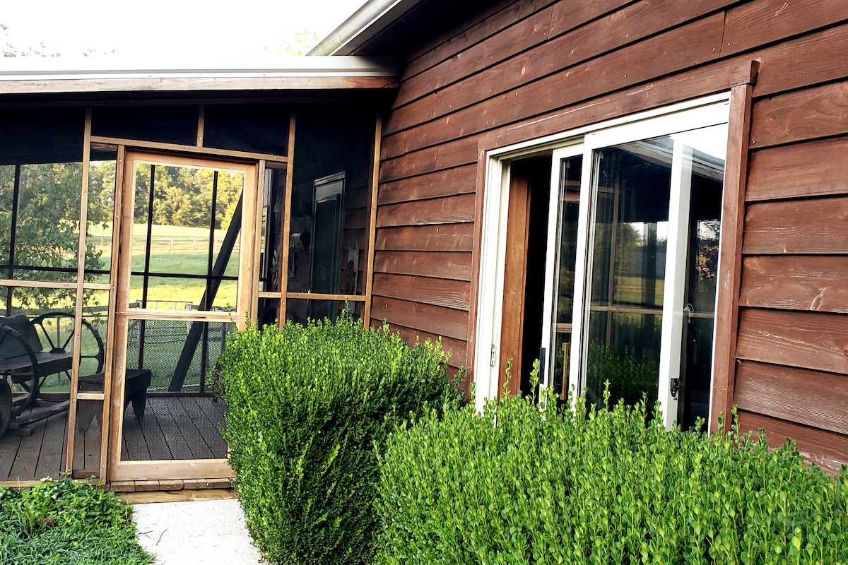 The exterior showcases a rustic cabin with wooden siding, complemented by neatly trimmed bushes. A screened porch is visible, providing a welcome entrance with wooden flooring and a partial view of the outdoor area beyond.