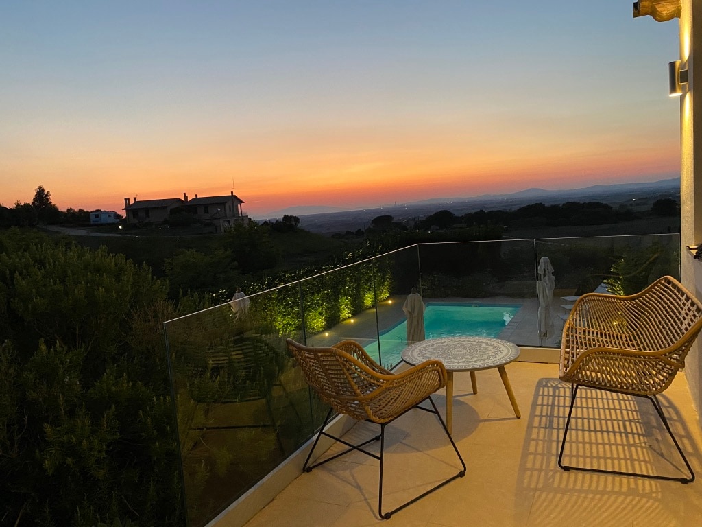 A serene terrace is shown at sunset, featuring two woven chairs and a round table. The glass railing allows for an unobstructed view of the garden and swimming pool, with distant hills and a glowing sky in warm hues creating a calming backdrop.