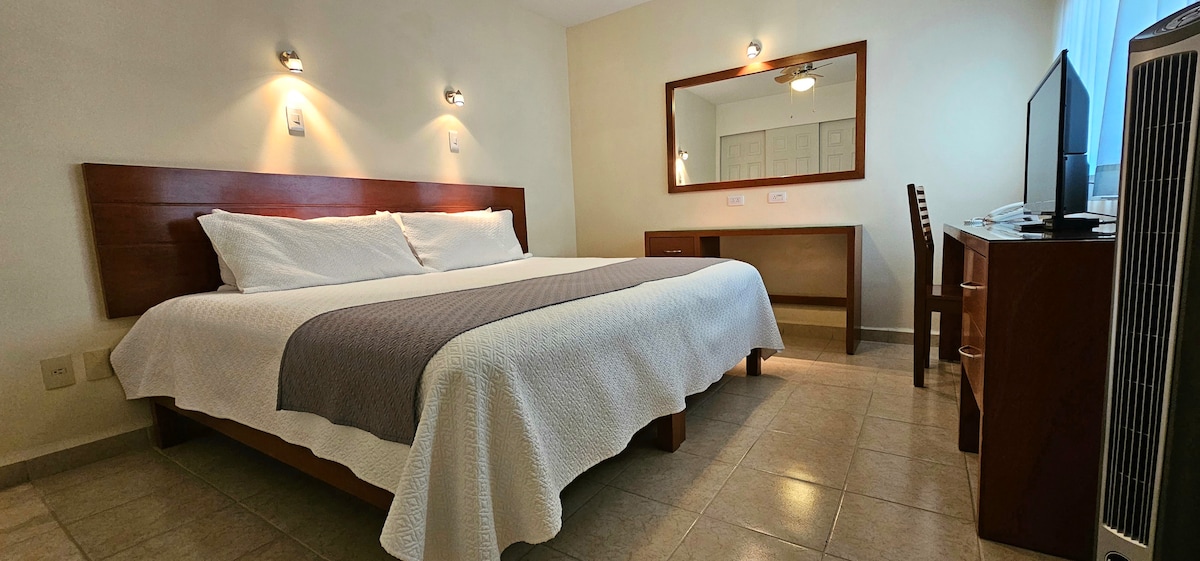 A cozy room features a spacious bed with crisp white bedding and a subtle gray accent blanket. A wooden desk and chair are positioned opposite a large mirror, while soft wall sconces provide gentle lighting. Natural light enters through a nearby window.