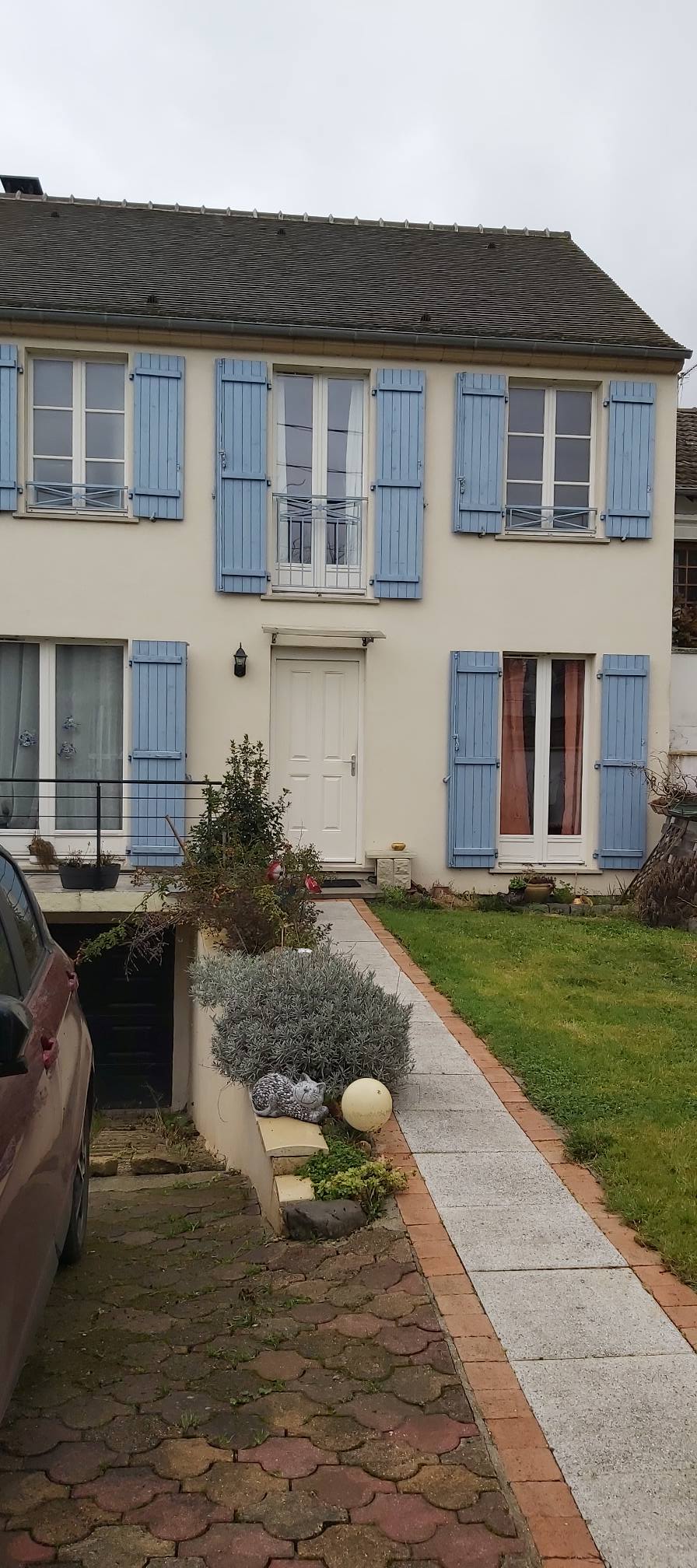 A charming two-story house is depicted with light blue shutters framing the windows. A neat pathway leads to the front door, bordered by a small garden featuring greenery and flowers. The exterior showcases a classic design with soft, muted colors.
