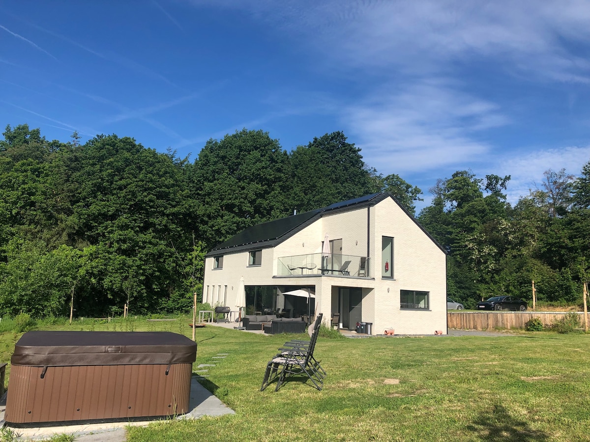 A modern vacation home is set against a backdrop of lush greenery. A spacious deck is visible with seating arranged for relaxation, while a hot tub is situated on the lawn, surrounded by an open area of well-maintained grass.