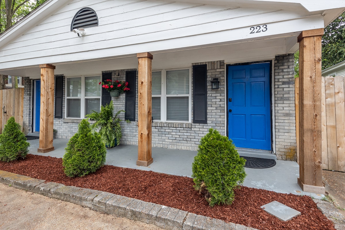 The exterior of a duplex is captured, featuring a bright blue front door framed by two wooden columns. Planters with flowers are positioned on the sides, and neatly trimmed shrubs are visible along the pathway. A gravel border defines the landscaped area.