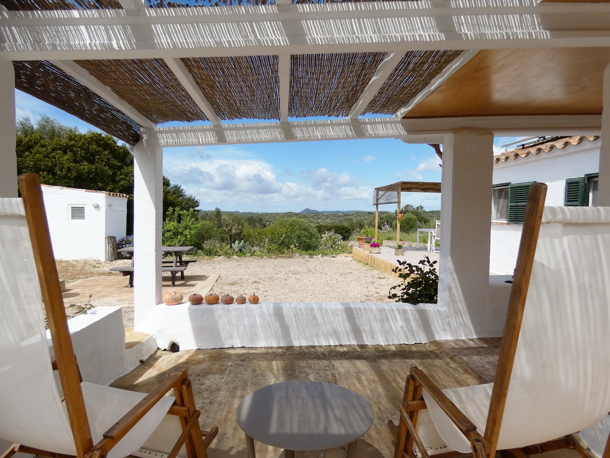 A shaded seating area features two white chairs and a small round table, overlooking scenic countryside views. The outdoor space is framed by wooden beams and natural textures, with a gravel pathway leading to vegetation and open fields in the background.