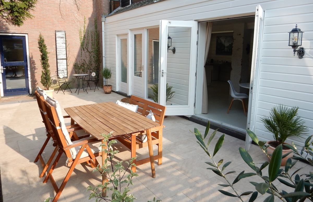 An outdoor dining area is featured, with a wooden table surrounded by matching chairs. French doors lead into the interior, and potted plants are positioned around the space, contributing to a relaxed garden atmosphere.