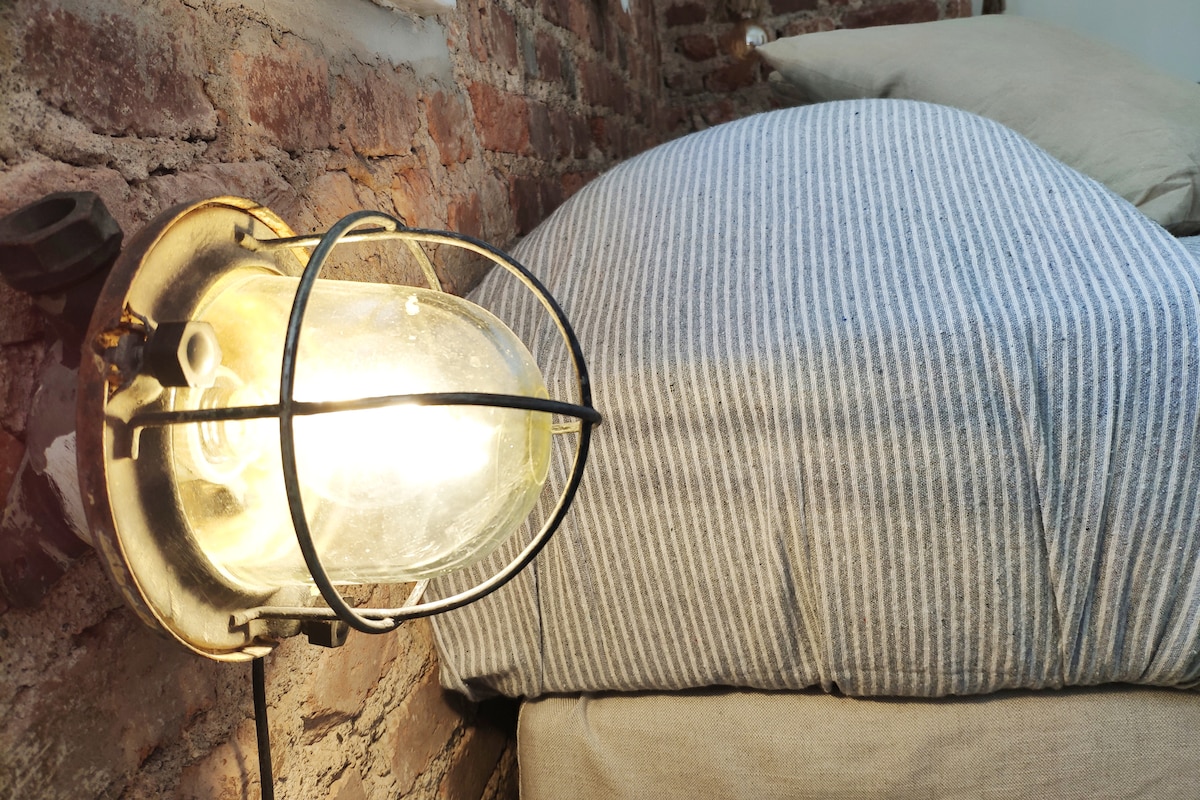 A bedside lamp with a protective metal cage emits a soft light, illuminating the area. A striped bed covering drapes over a spacious mattress, which is positioned against a rough brick wall, adding texture to the room.