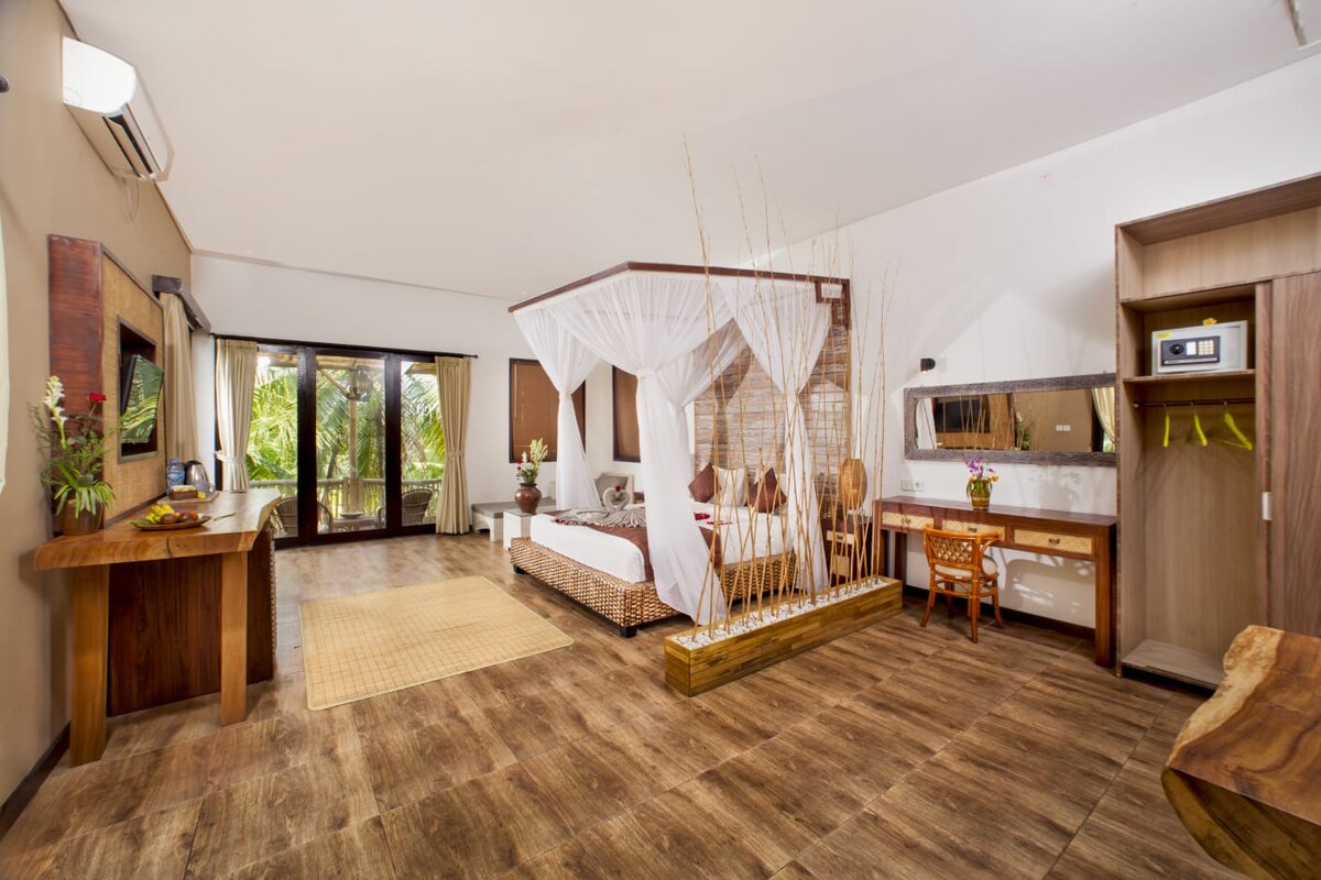 The spacious room features a four-poster bed with sheer drapery and is surrounded by natural light from sliding glass doors. Wooden accents and tropical décor create a soothing ambiance. A comfortable seating area and a well-appointed dresser are included, enhancing the overall comfort.