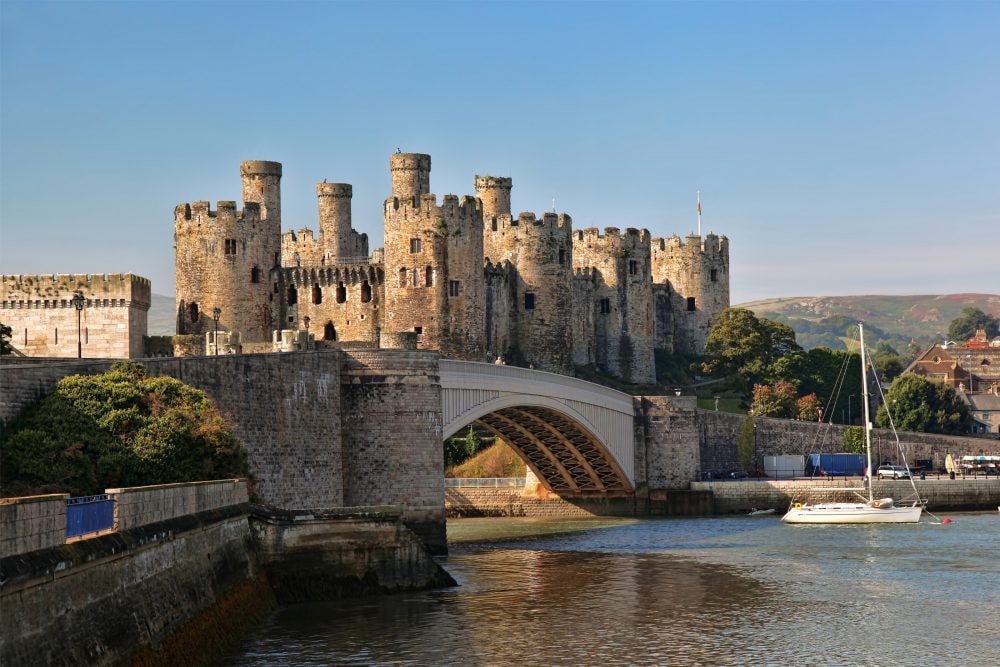 A historic castle with numerous towers and battlements is seen rising above the water, reflecting a mix of stone and scenic greenery. A bridge leads to its entrance, while a sailboat is anchored nearby, complementing the tranquil river view.