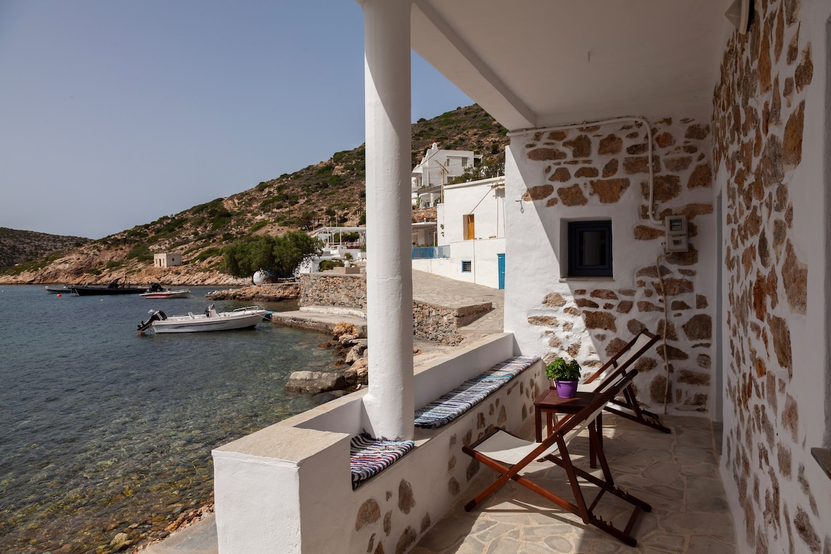 A tranquil porch is situated just steps from the sea, featuring two folding chairs and a small table. The view includes clear water and nearby boats, while the surrounding hillside showcases traditional structures and greenery.