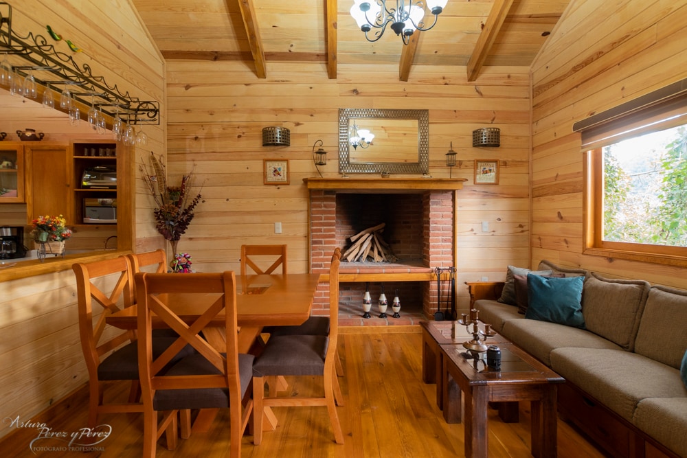 The interior of the cabin features warm wooden walls and ceiling beams. A dining table with x-back chairs is present, beside a cozy seating area with a sofa. A fireplace is positioned at one end, with logs neatly stacked for use. Natural light enters through adjacent windows.