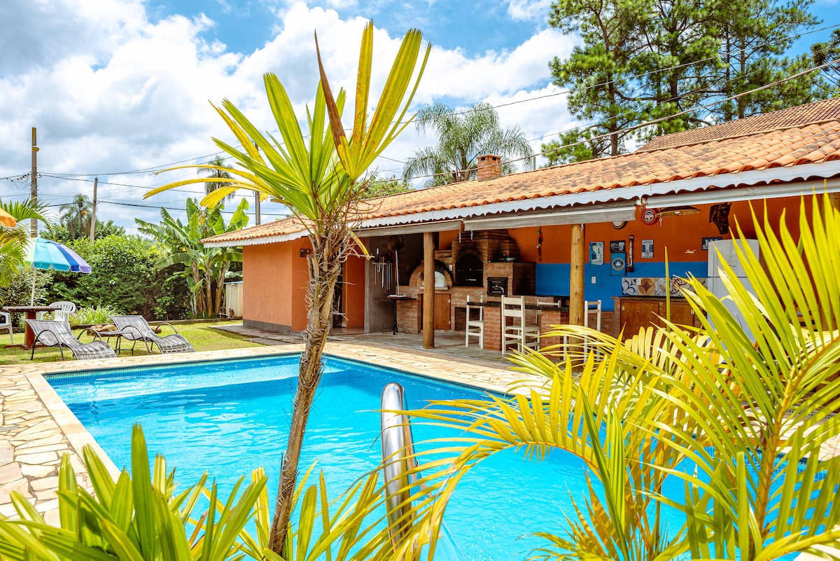The image showcases a calm outdoor space featuring a turquoise pool surrounded by vibrant green plants. Lounge chairs are positioned beside the pool, and a shaded area with a barbecue grill is visible in the background under a tiled roof.