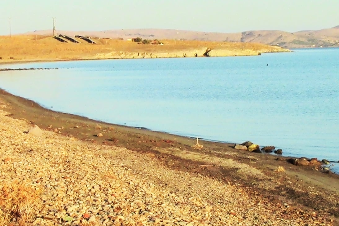 A tranquil seaside view showcases a gentle curve of a sandy beach bordered by smooth pebbles. The calm water reflects the light of the sun, creating a serene atmosphere. Distant hills are visible in the background, adding to the natural landscape.