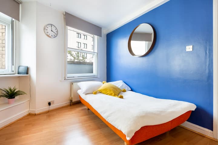 Camera luminosa con divano letto pieghevole, parete con accenti blu e grandi finestre che lasciano entrare molta luce.